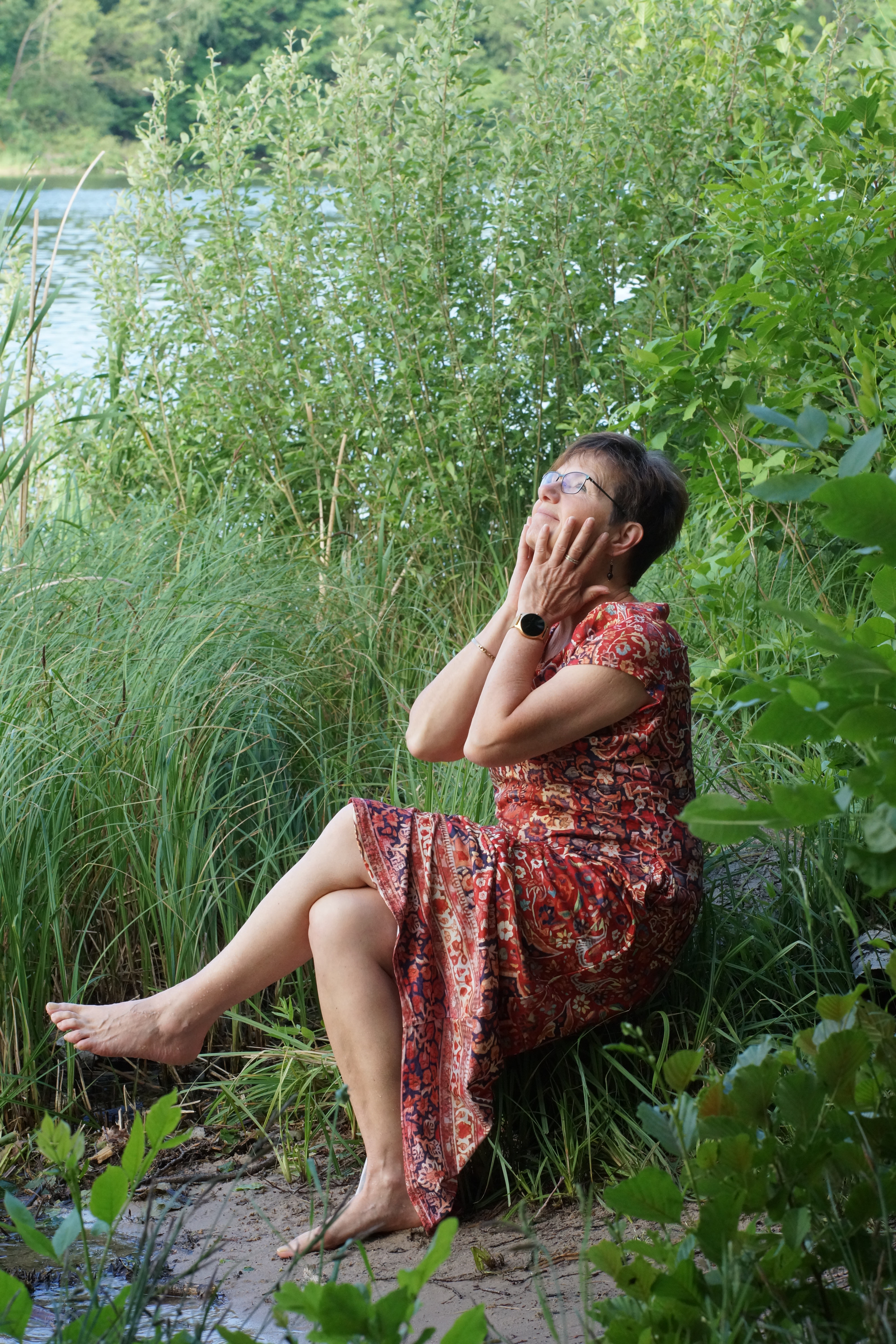 Frauenfotografie Frau  in rotem Kleid genießt es am Wasser zu sitzen und streicht sich über ihr Gesicht