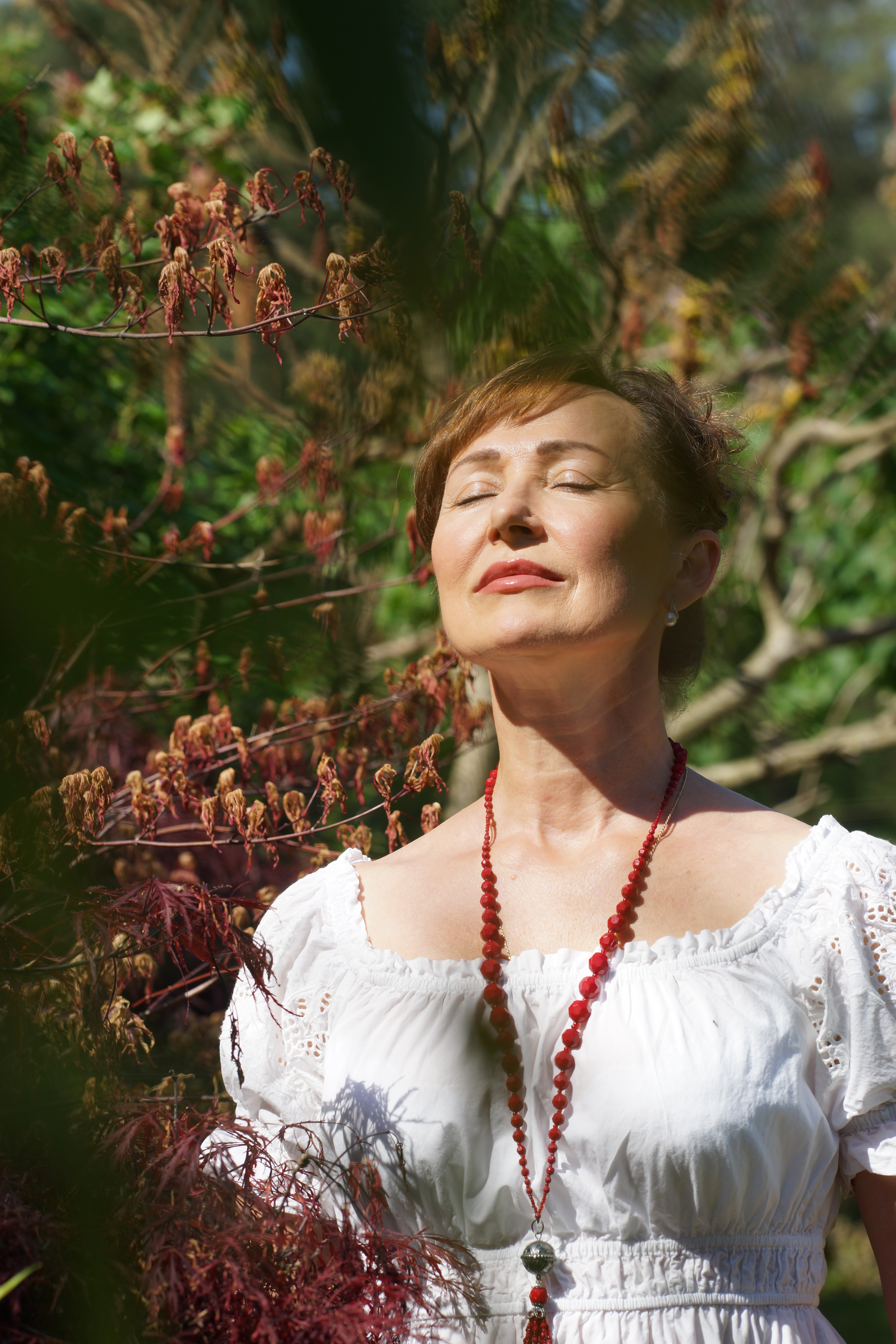 Frauenfotografie - brünette Frau in weißem Oberteil mit roter Kette  hält ihr Gesicht mit geschlossenen Augen ins Licht und ist umrahmt von roten Blättern